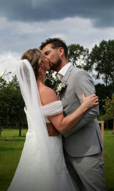 Een pasgetrouwd stel kust elkaar in een groene omgeving onder een bewolkte lucht.
