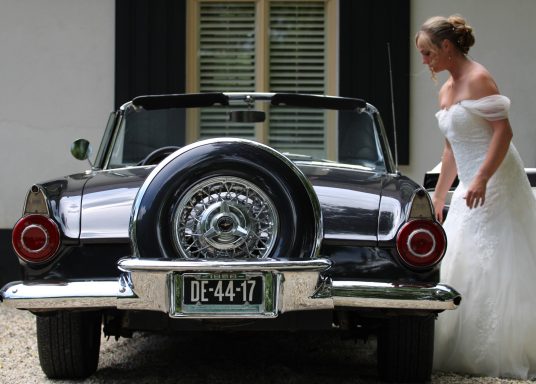 Bruidsmeisje in een witte jurk naast een vintage auto.