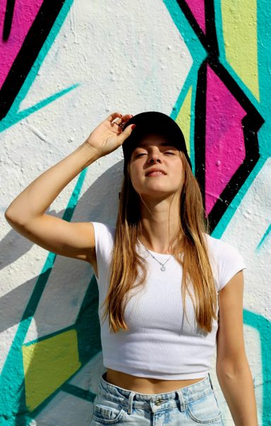 Jonge vrouw in witte top en pet poseert voor een kleurrijke muurschildering.