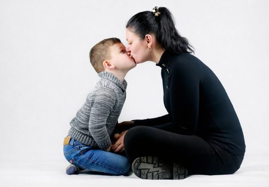 Moeder en kind delen een kus, zittend op de grond tegen een witte achtergrond.