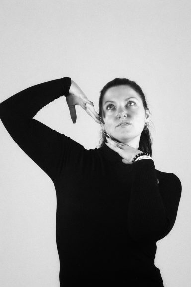 Vrouw in zwarte kleding staat met arm omhoog en kijkt nadenkend omhoog.